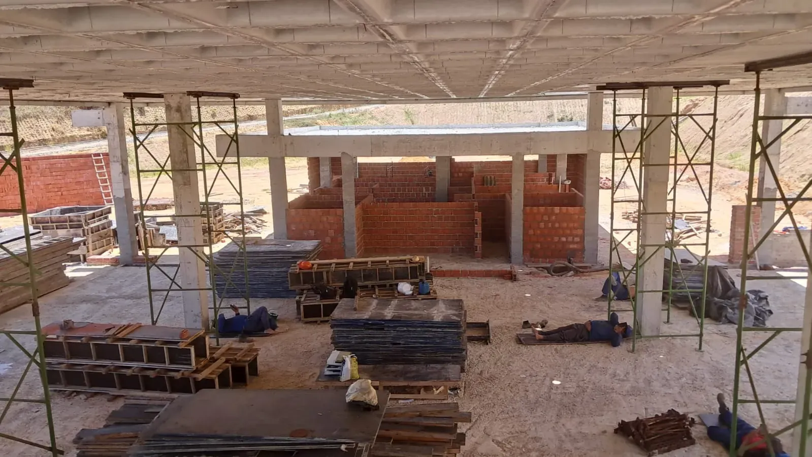 Sob a laje, os ambientes internos de alvenaria tomando forma, entre as torres de andaime e as fôrmas empilhadas.