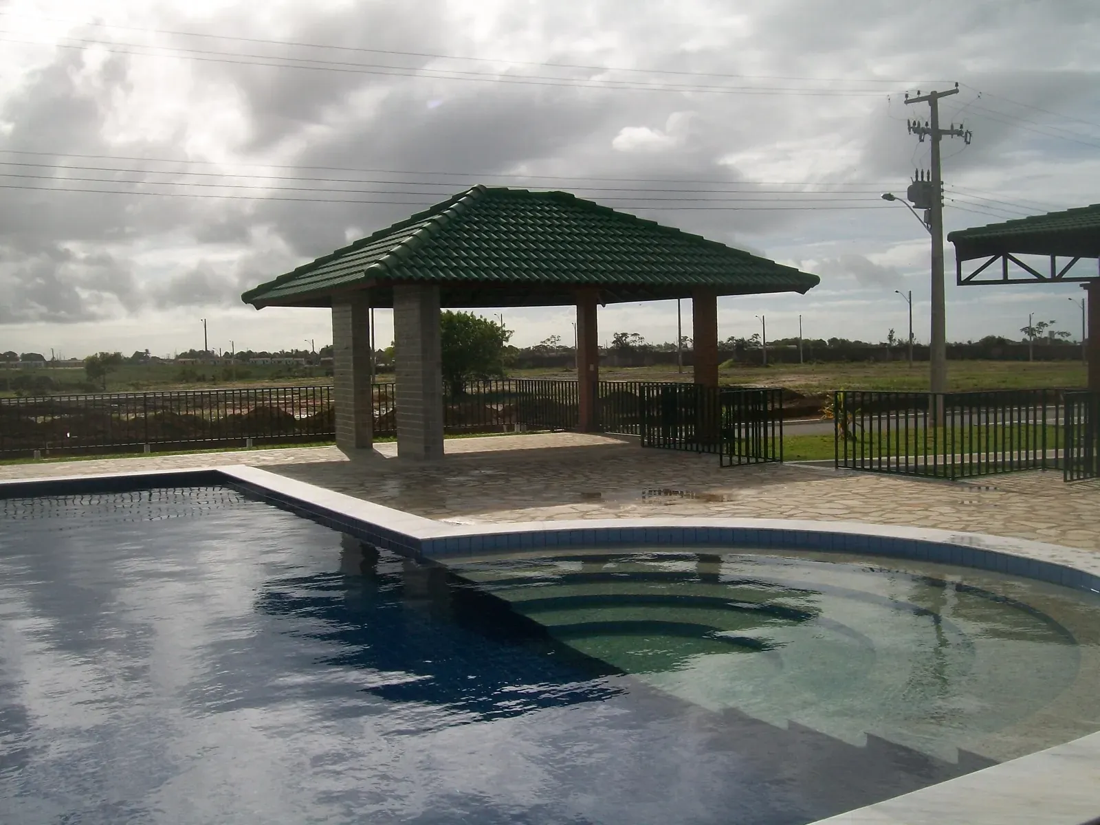 Piscina com deck de pedra e quiosque de telhado verde, o loteamento ao fundo