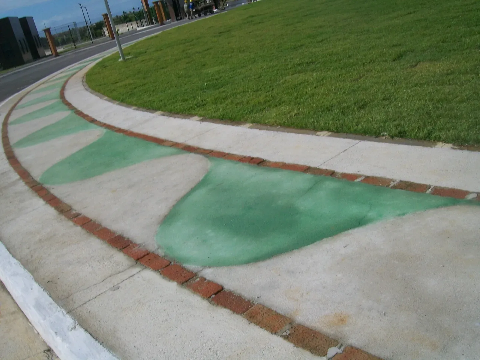 Pista de caminhada em concreto com faixa verde e borda de tijolo, contornando o gramado