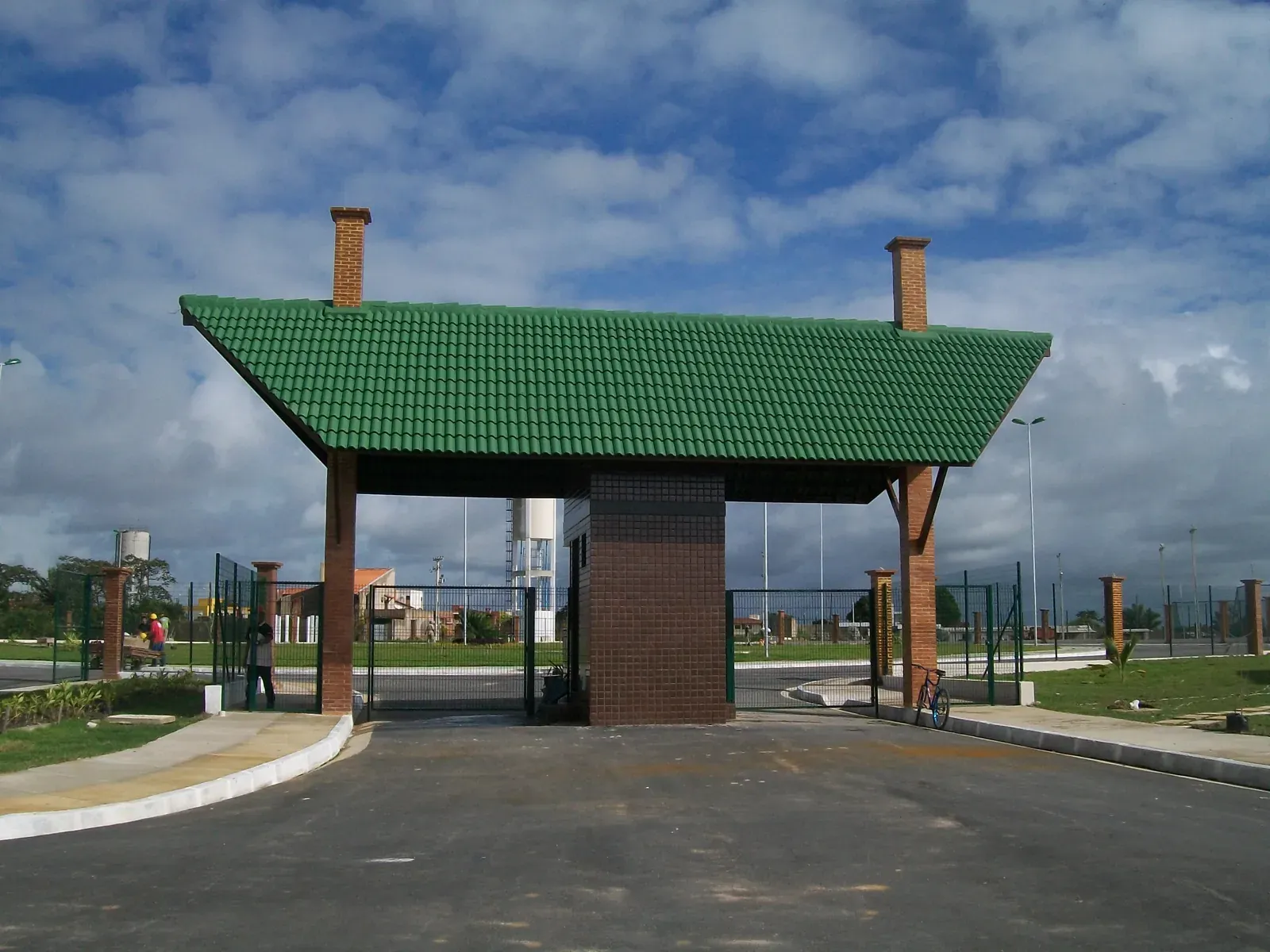 Pórtico de entrada do Reserva Bella Vista, com telhado verde sobre pilares de tijolo aparente e a área de lazer ao fundo