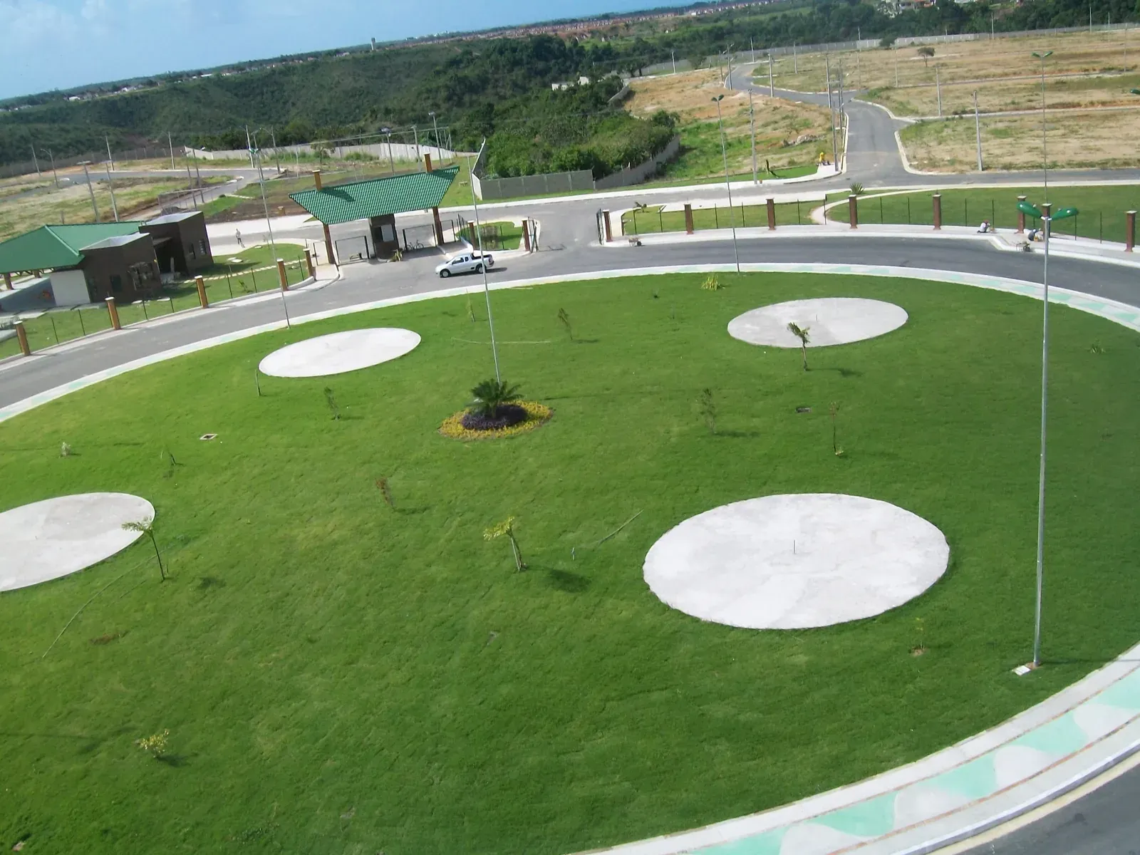 Praça circular gramada com quatro rodas de concreto e árvores recém-plantadas