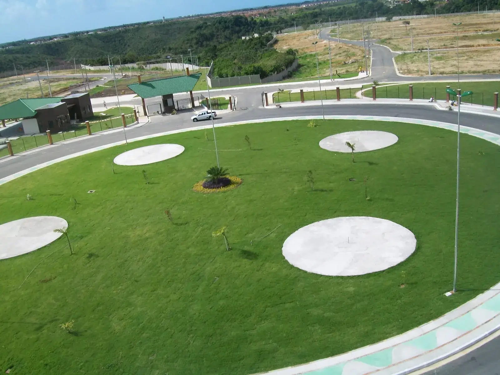 Praça circular gramada com quatro rodas de concreto e árvores recém-plantadas