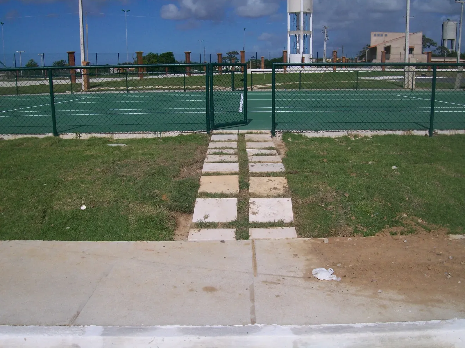 Quadra de tênis com piso verde e alambrado, com o reservatório de água ao fundo