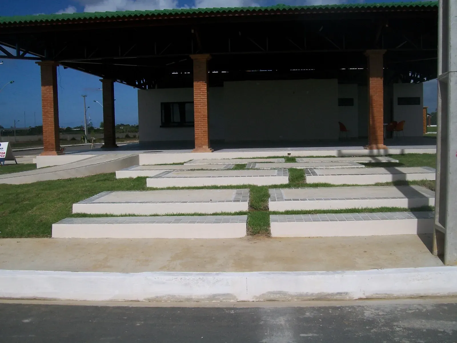 Salão de festas coberto, com pilares de tijolo aparente e passeio de placas de concreto entre gramado